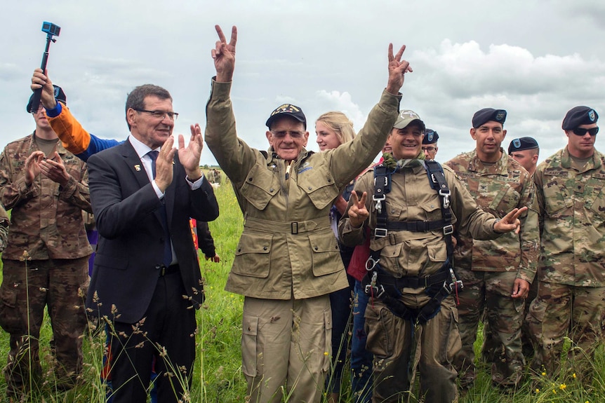 97yo in army fatigues holds two victory signs in the air surrounded by other parachutists in a field.