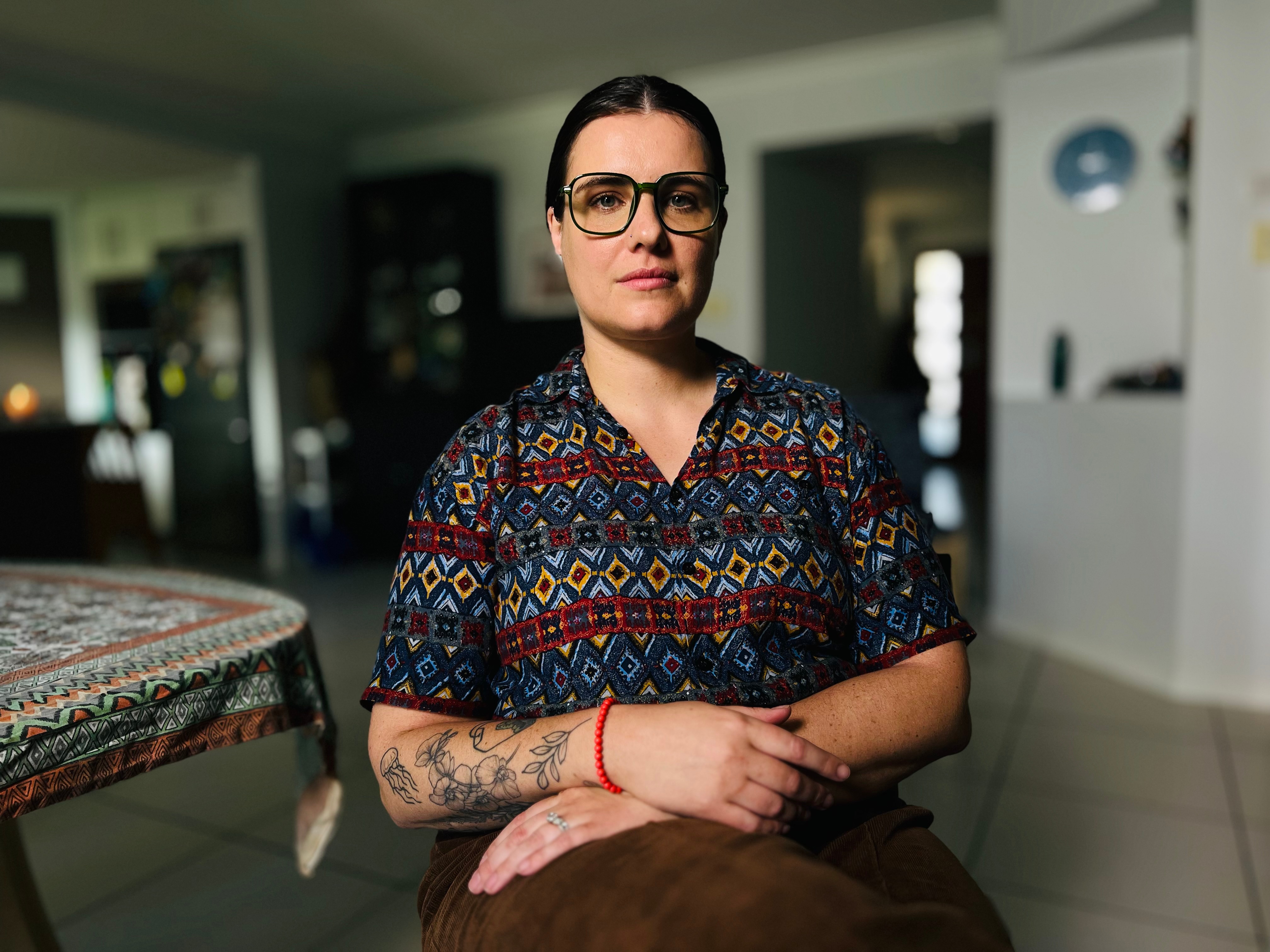 a young woman wearing a dark patterned top and glasses inside a suburban home
