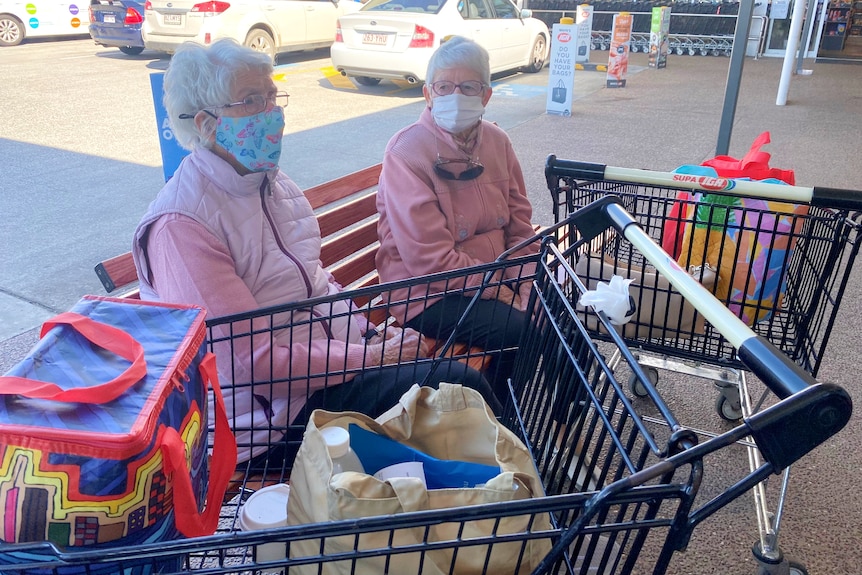 Two older women sit on a bench at a shopping complex with trollies containing groceries. They have masks on