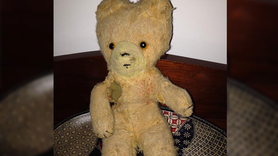A very worn teddy bear sits on a bed.