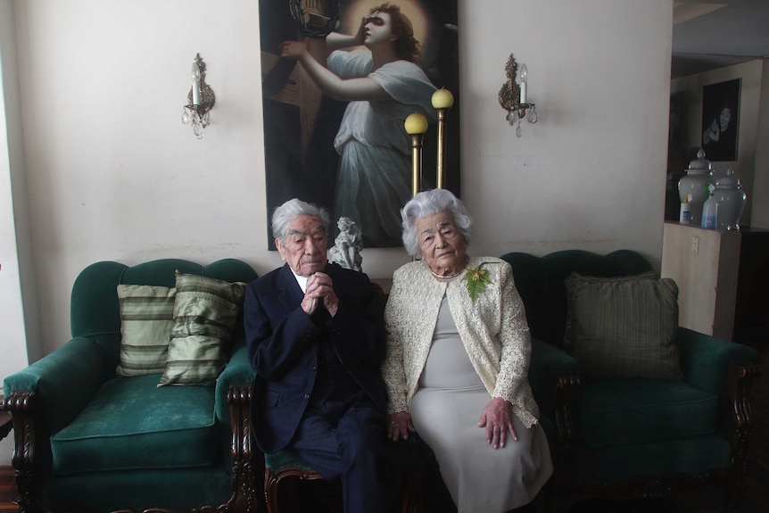 An old couple, man wearing a suit and a woman an ivory dress, sit on a couch.