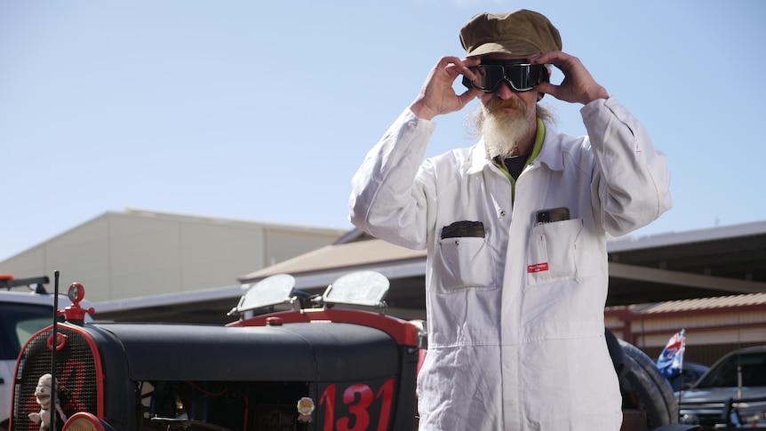 A man in a white jumpsuit and driving goggles stands in front of a vintage car.