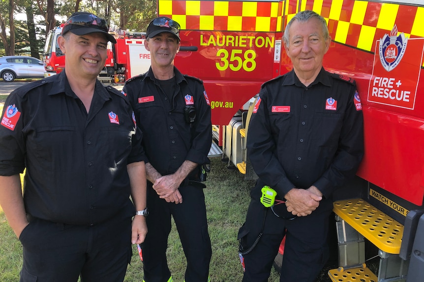 Three male firefighters in a dark uniform stand next to a fire truck marked 'Laurieton 358'.