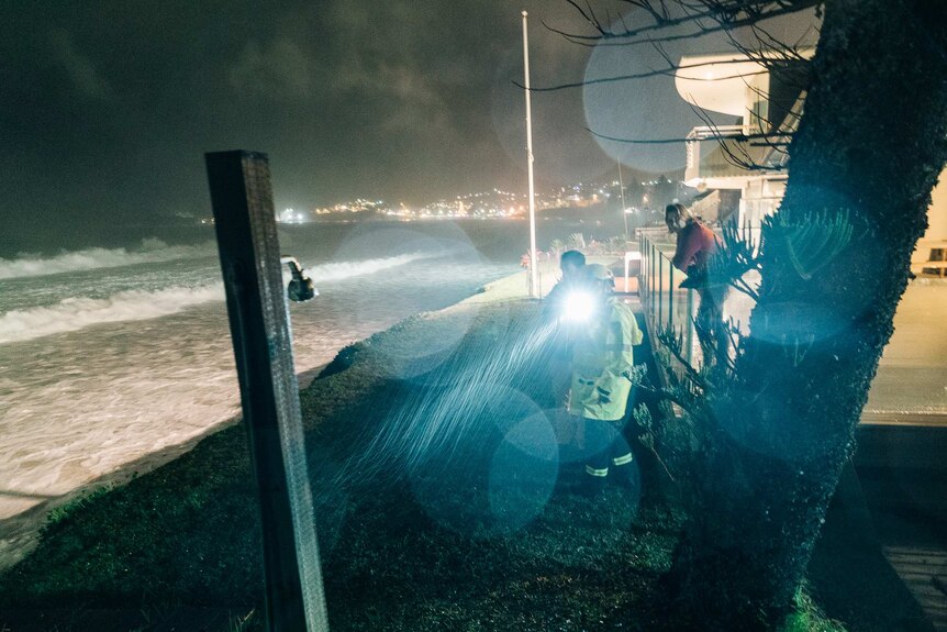 A firefighter points a torch in the rain at night on the edge of a cliff at Wamberal Beach with two residents looking on.