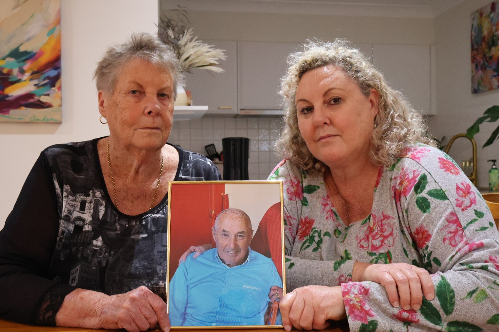 Two women looking sad holding a framed photo of a man.