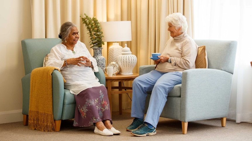 Two older women in aged care sit in armchairs talking and drinking tea.