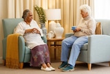 Two older women in aged care sit in armchairs talking and drinking tea.