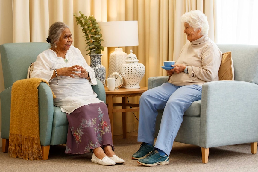 Two older women in aged care sit in armchairs talking and drinking tea.