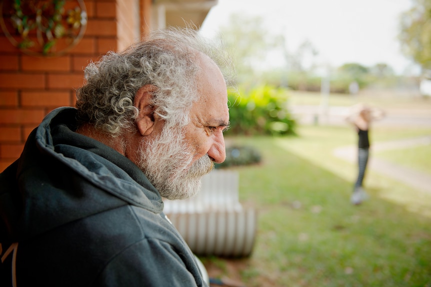 A man wearing a hooded jumper sits on a chair outside his unit and looks to the ground. 