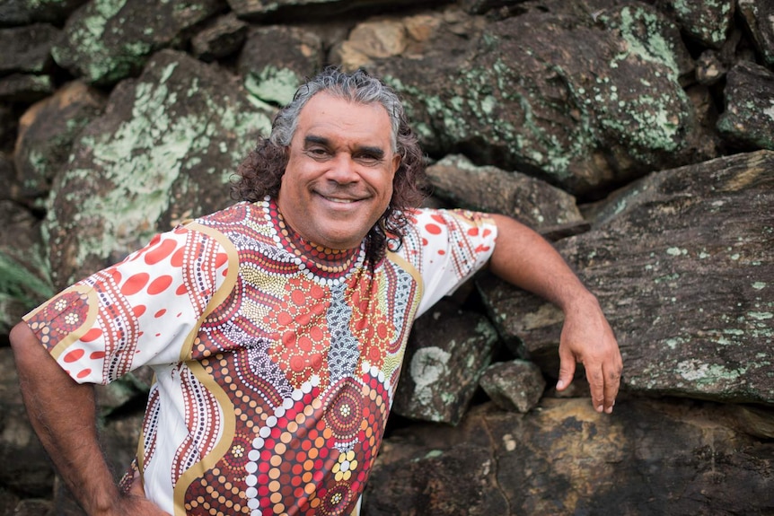 An Indigenous man with curly, shoulder-length hair, wearing a bright Indigenous print t-shirt, leaning against a rock wall.