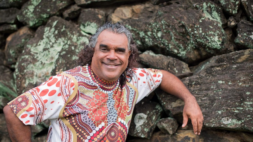 An Indigenous man with curly, shoulder-length hair, wearing a bright Indigenous print t-shirt, leaning against a rock wall.