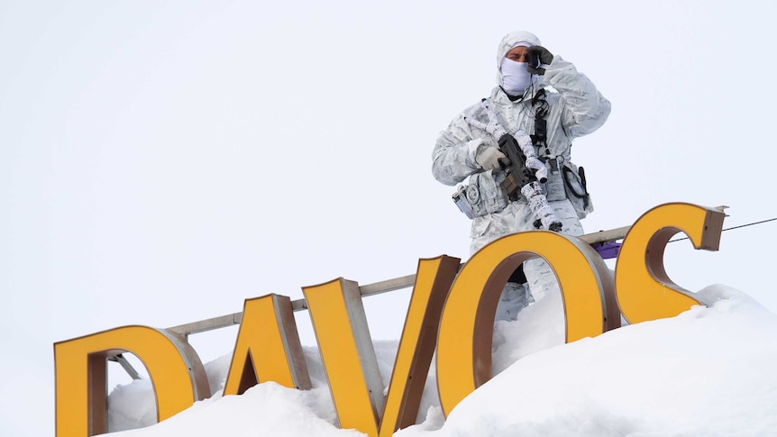 An armed Swiss police officer stands guard on the snowy roof of a hotel next to a large yellow sign which says Davos