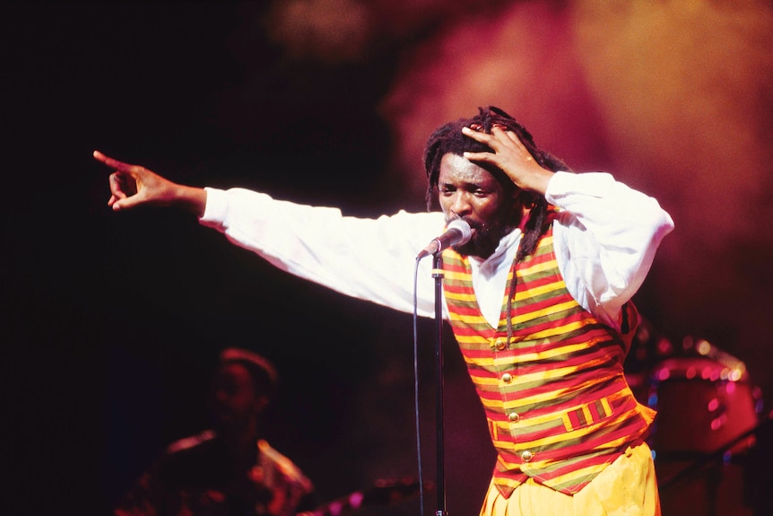 A musician with dreadlocks and a vest sings into a microphone onstage.