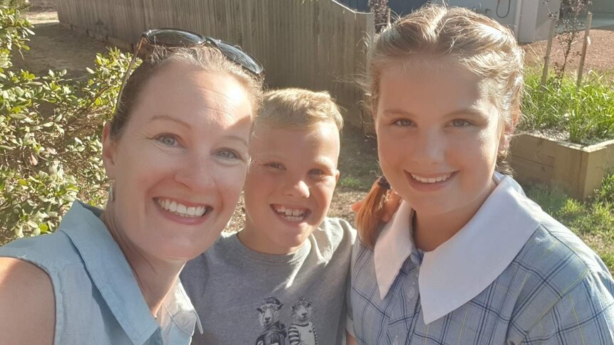 A smiling mother stands with her two young children as they look into the camera.