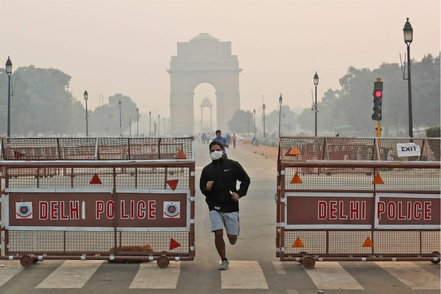 A man wearing a mask runs through a barrier that says DELHI POLICE. India Gate is visible in the background, the air is smoggy