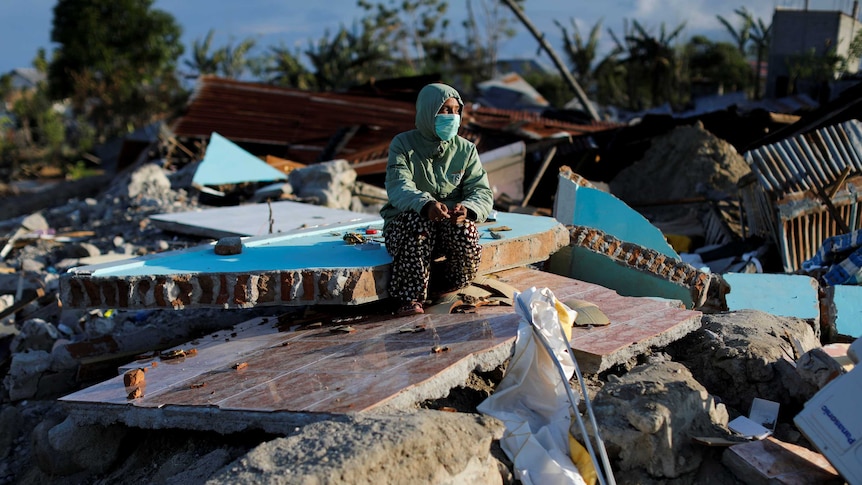 A woman in a hoodie and face mask crouches on rubble