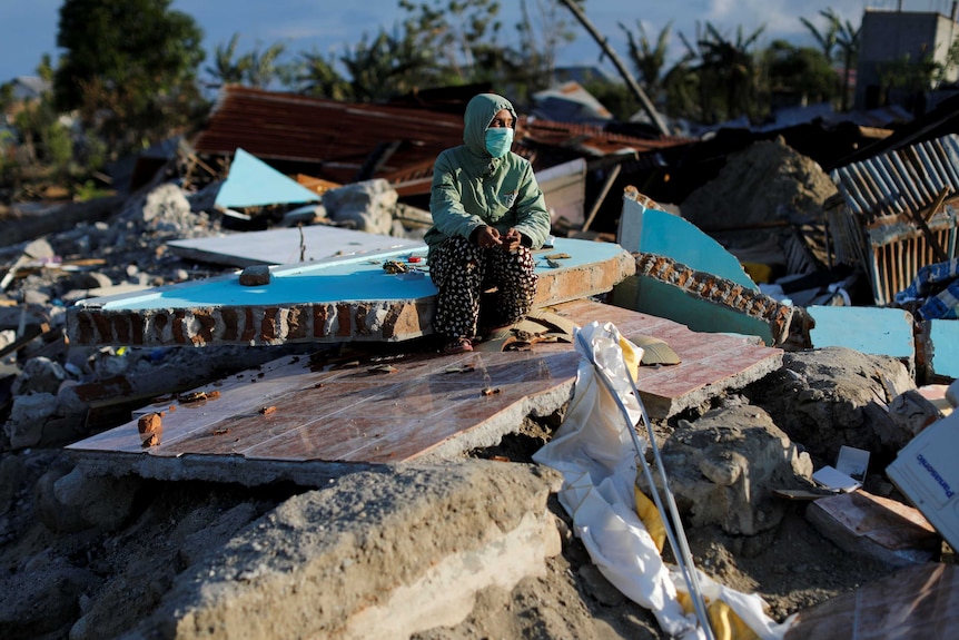 A woman in a hoodie and face mask crouches on rubble