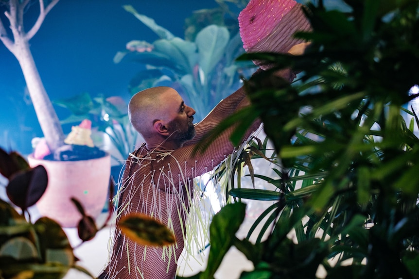 A bald man in a mesh shirt holds a basket upside down. He is standing on stage, and can be seen past the leaves of plants.