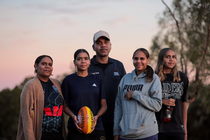 Four Aboriginal women and one young man stand smiling beneath pinkish sunset with trees in background