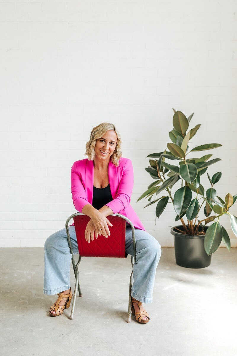 Portrait of Jill Start in a pink blazer and jeans sitting backwards on a chair, a rubber fig plant in the background.