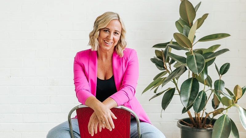 Portrait of Jill Start in a pink blazer and jeans sitting backwards on a chair, a rubber fig plant in the background.