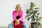Portrait of Jill Start in a pink blazer and jeans sitting backwards on a chair, a rubber fig plant in the background.
