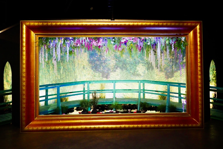 Darkened gallery space with large golden frame around a lily pond scene, with a walkway running through picture.