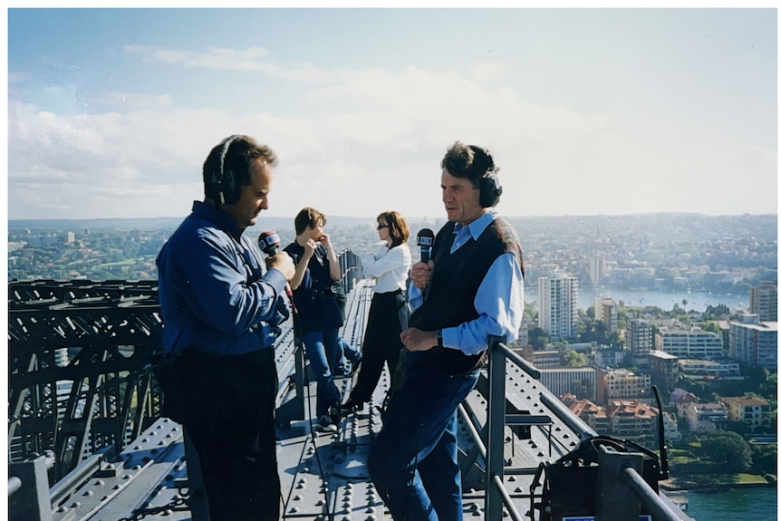 A man holds a microphone and wears headphones as he talks to another man with a view of Sydney below them. 