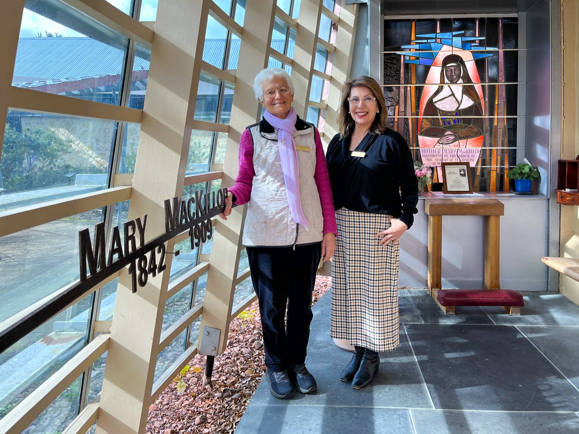 Two women standing with a stained glass window of a nun wearing a habit