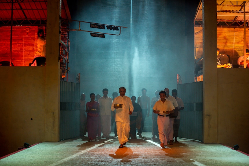 A group of people in white kurtas emerge onto a smoky stage, holding offerings, with musicians playing in the wings.