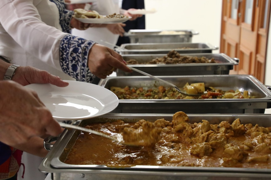 Hands of people ladelling in curry and meat onto white plates from Bain Marie