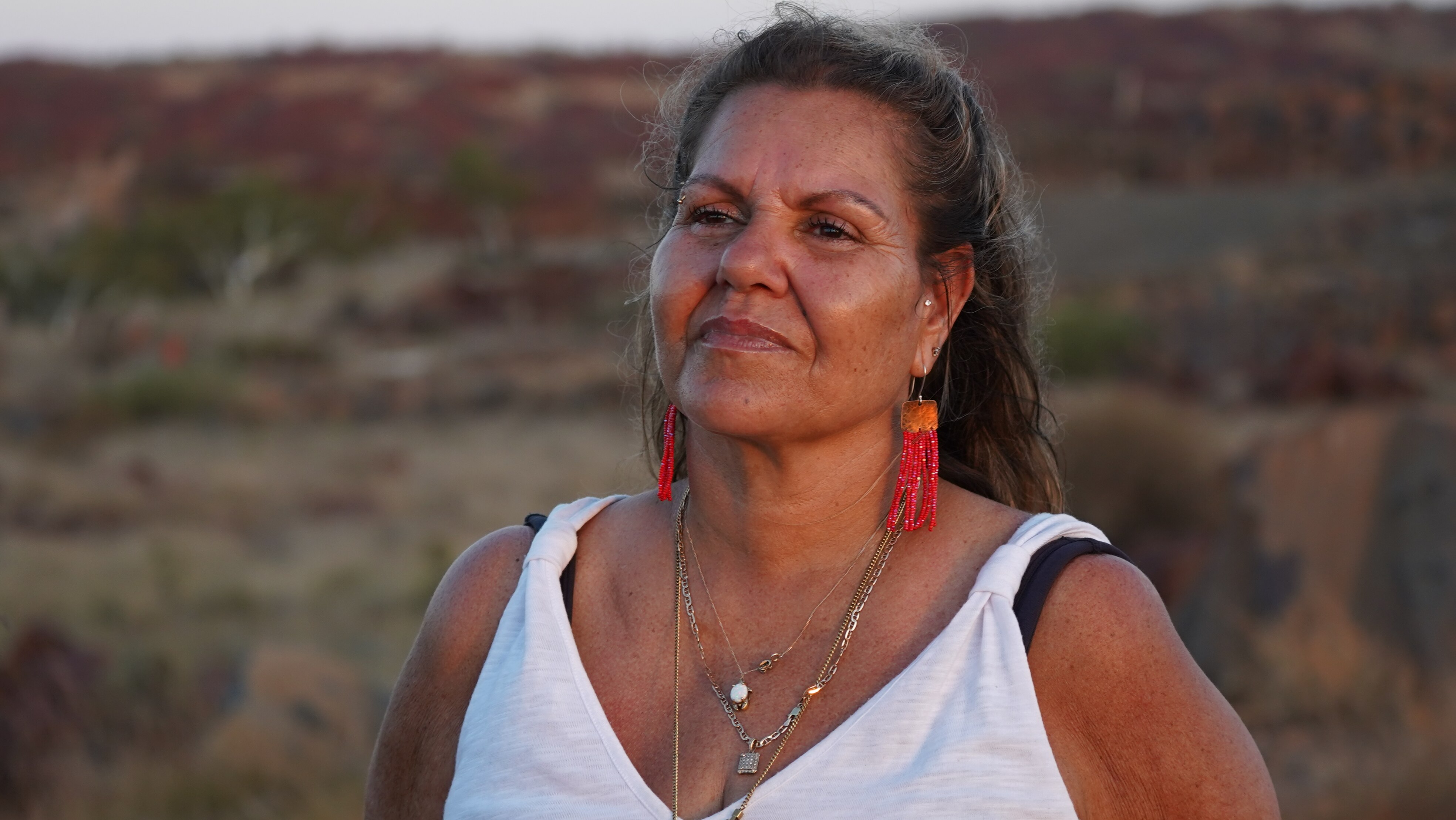Una mujer bronceada que llevaba una camiseta blanca y un arete rojo contra una llanura rocosa y seca al atardecer.