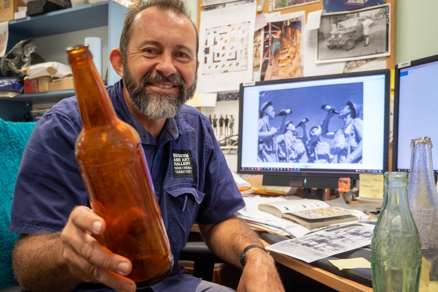 Man in busy office at computer desk with brown bottle.