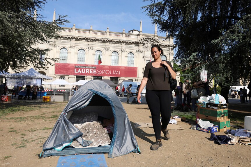 A woman stands next to a tent which sits of dirt outside of a large ornate building in Milan, Italy.