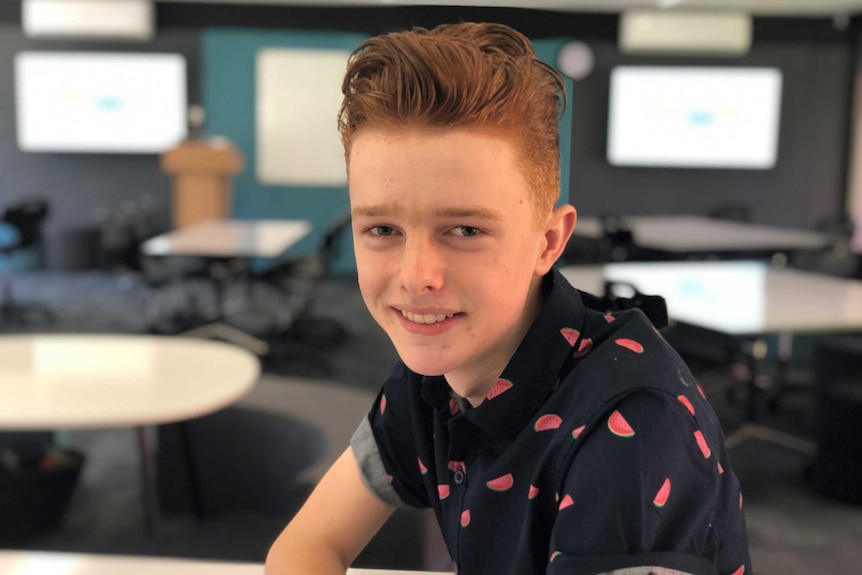 A teenager wearing a shirt with watermelons on it sits in a classroom and smiles for the camera