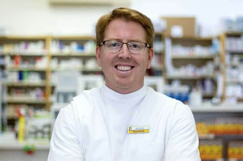 A man wearing a pharmacist uniform stands in front of shelves of medicine.