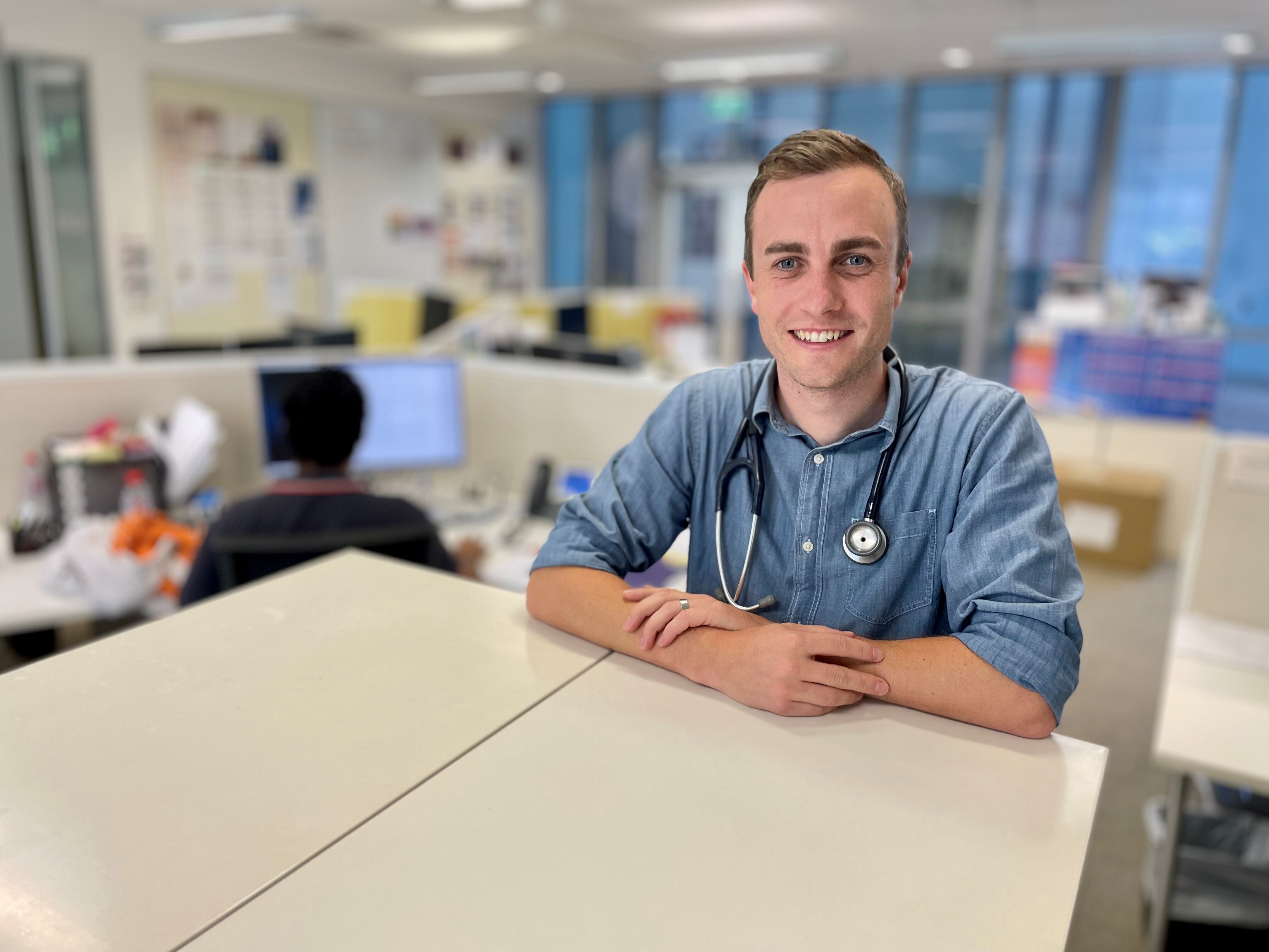 A young doctor stands in an office leaning against a table.