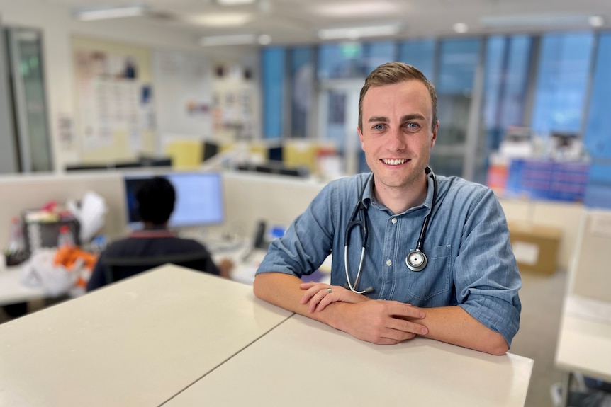 A young doctor stands in an office leaning against a table.