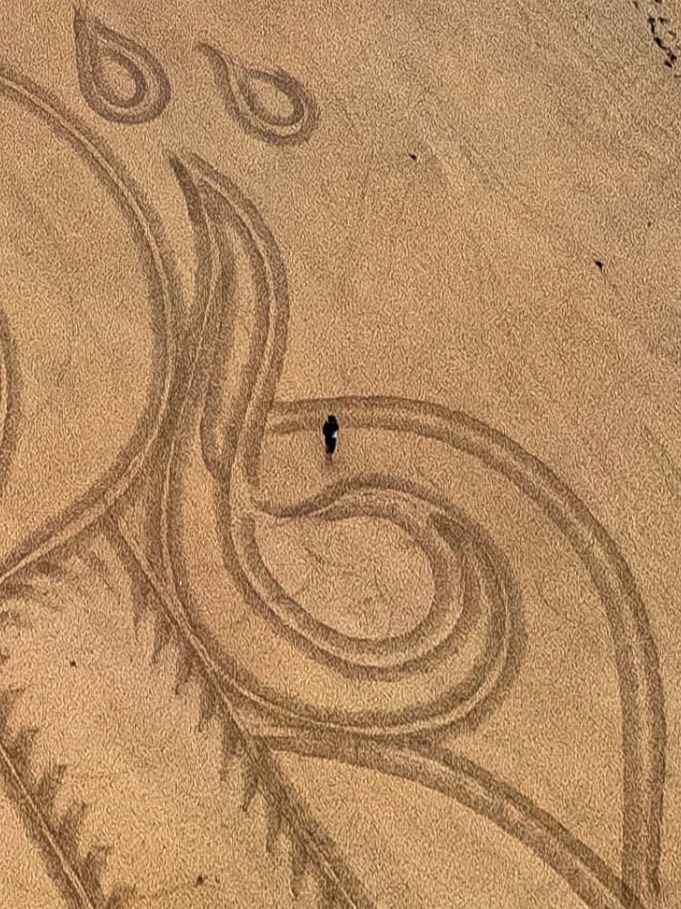 A drone shot from above, a man stands in the artwork he's drawn on the sand.
