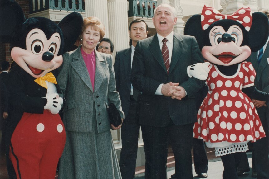 Mikhail Gorbachev dressedin his suit and a woman dressed in grey paints and a jacket pose next to Mickey and Minnie Mouse.