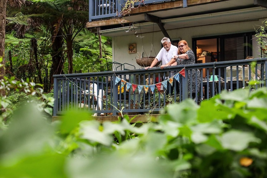 A man and a woman stand on their front balcony, adorned with flags and hanging plants. The woman is pointing at her garden.