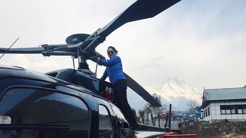 Woman wearing a thick jacket and aviators stands on the side of a helicopter.
