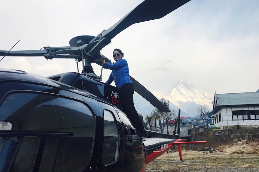 Woman wearing a thick jacket and aviators stands on the side of a helicopter.