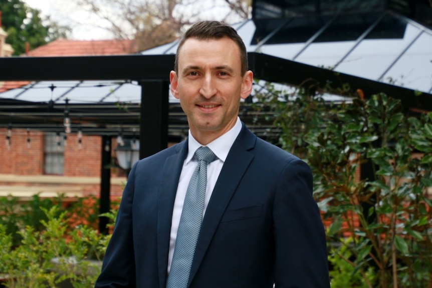 A man wearing a navy blue suit and tie stands in front of a garden bed.