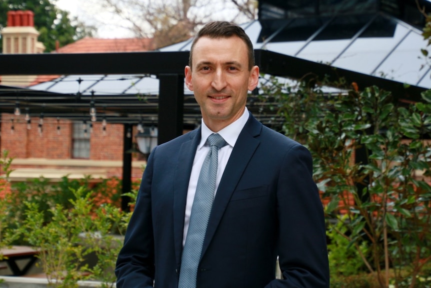 A man wearing a navy blue suit and tie stands in front of a garden bed.