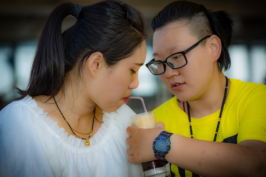 A woman holds a drink as her partner sips from the straw.