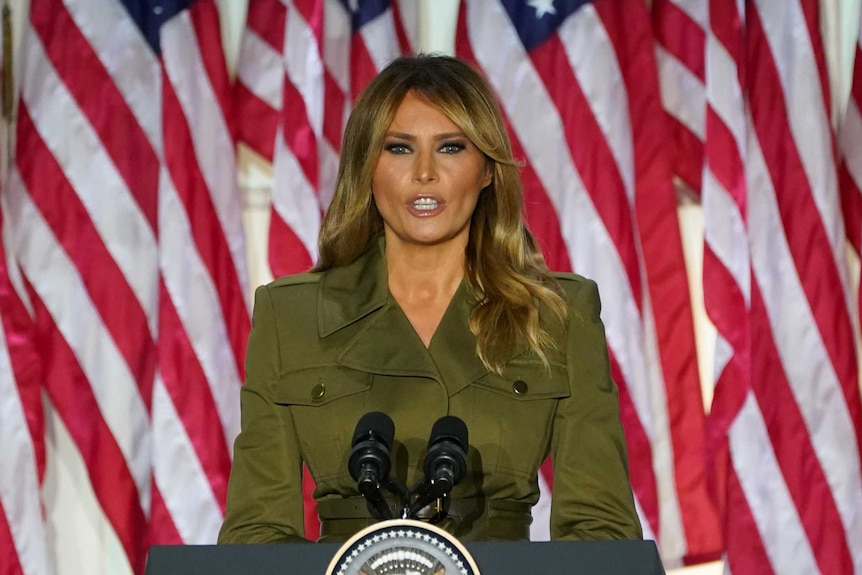 Melania Trump in an olive suit speaking at a lectern behind a row of American flags