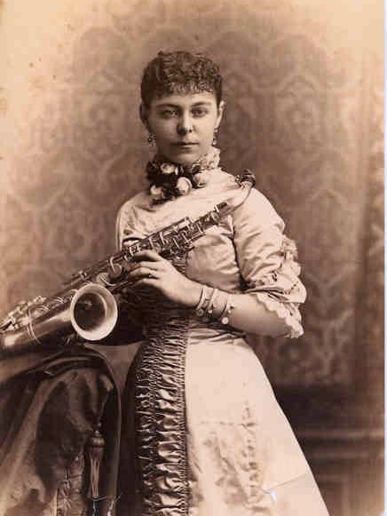 Black and white photo of woman in dress with floral scarf at her neck, holding a saxophone. She looks ahead with closed mouth.