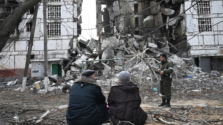 An elderly couple sits on a log opposite a residential building destroyed by a missle strike.