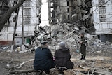 An elderly couple sits on a log opposite a residential building destroyed by a missle strike.